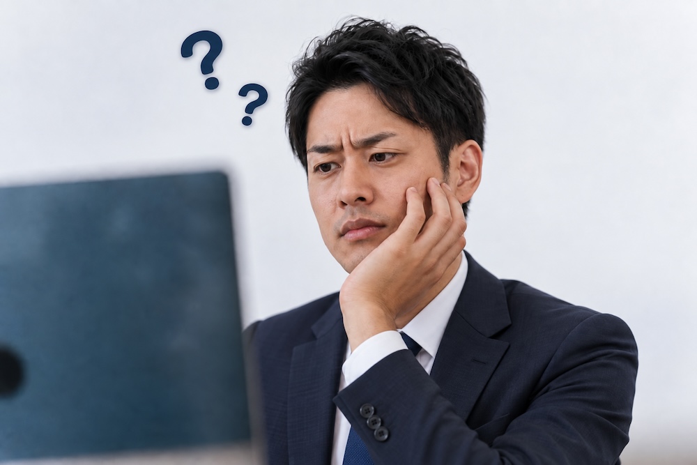 パソコンの前で悩むスーツ姿の男性の画像。